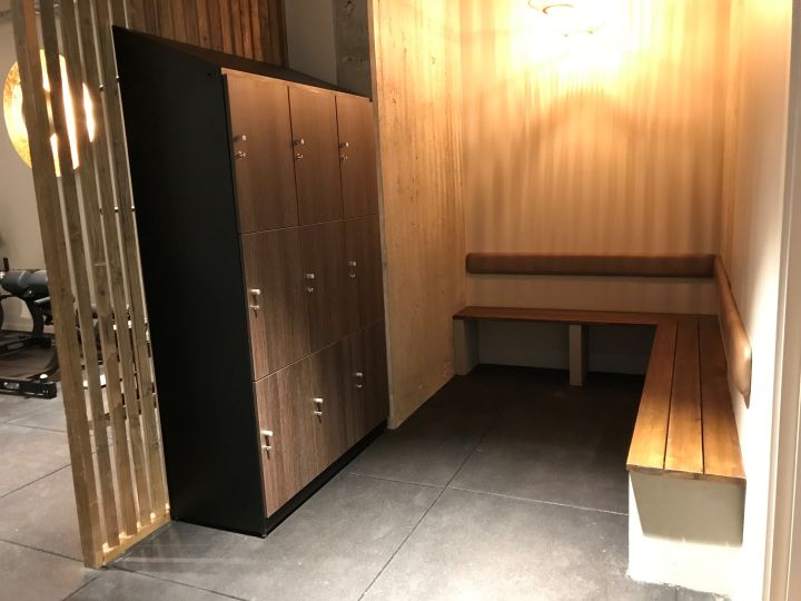 Træningscenters omklædningsrum med skabe, bænk og håndvægte i baggrunden til kommercielt fitnessmiljø